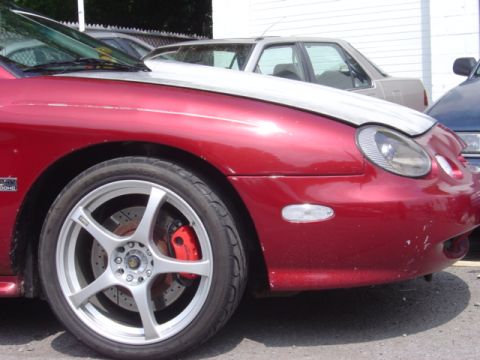 1998 Ford Taurus - Close-up of a red sedan's front fender, wheel, and part of the hood, with other vehicles in the background.