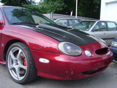 1998 Ford Taurus - Front view of a red 1998 Ford Taurus with a black hood, parked outdoors with other vehicles.