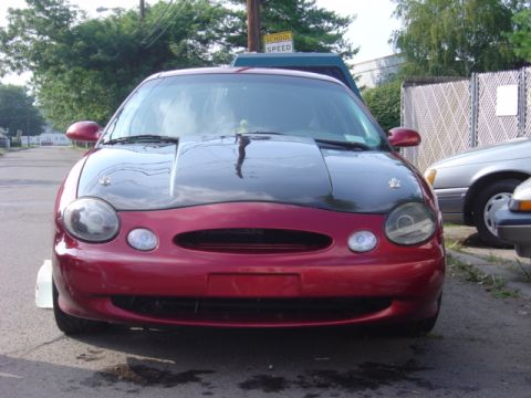 1998 Ford Taurus - A red 1998 Ford Taurus with a black hood is parked on a street, with another vehicle visible to the right.