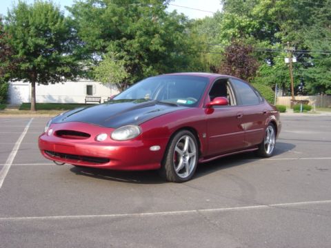 1998 Ford Taurus - A red sedan with a black hood is parked in an empty lot on a sunny day.