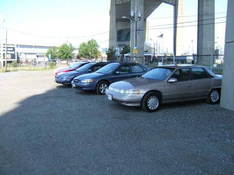 1998 Ford Taurus - A row of sedans are parked in a lot under a bridge, with the closest vehicle being a tan sedan.