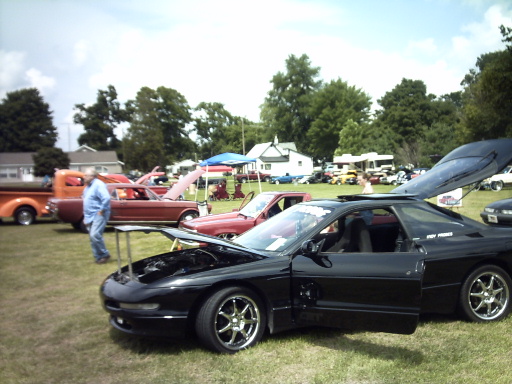 Black sports car with hood and trunk open at an outdoor car show. Other cars and people are visible in the background