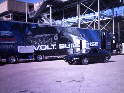 1995 Ford Probe - A black 1995 Ford Probe is parked beside a large Ford racing trailer at an event.