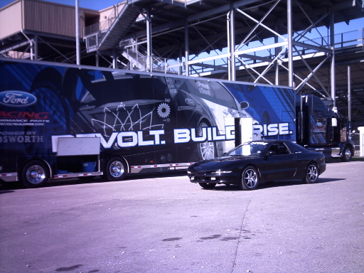 Black 1995 Ford Probe parked next to a large Ford racing trailer with "VOLT. BUILD. RISE." text