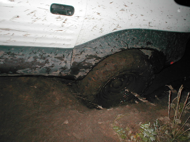 White SUV with mud-splattered side and rear wheel stuck deep in mud at night