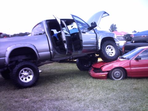 1995 Ford Probe - A lifted pickup truck is parked on top of a red car, causing significant damage to the car's front.