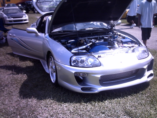 Silver Toyota Supra with hood and driver door open, aftermarket body kit. Another car visible in background