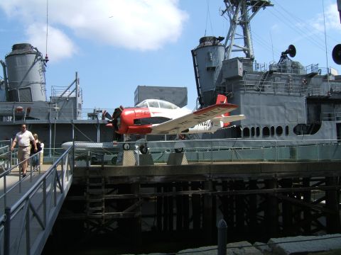 1998 Ford Taurus - A vintage propeller plane is displayed on the deck of a large gray naval vessel under a blue sky.