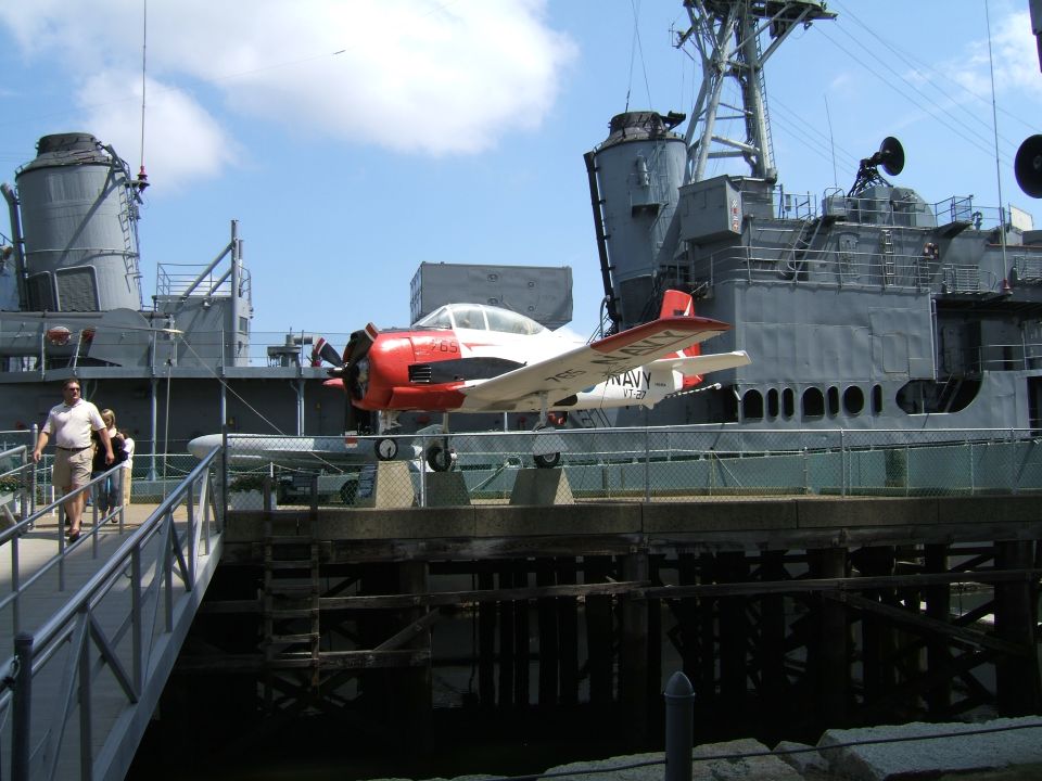 Red and white propeller airplane with NAVY markings on a gray naval ship