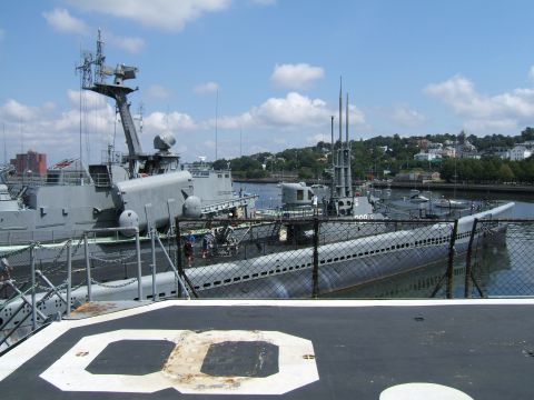 1998 Ford Taurus - A view of docked submarines and a naval ship against a backdrop of a coastal town under a partly cloudy sky.