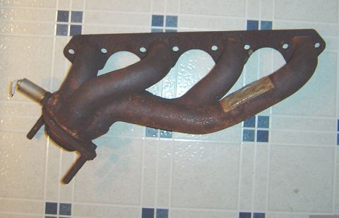 1986 Ford Bronco - A rusty four-port cast iron exhaust manifold with tubular runners laid on a white and blue tile floor. An oxygen sensor bung with a stainle…