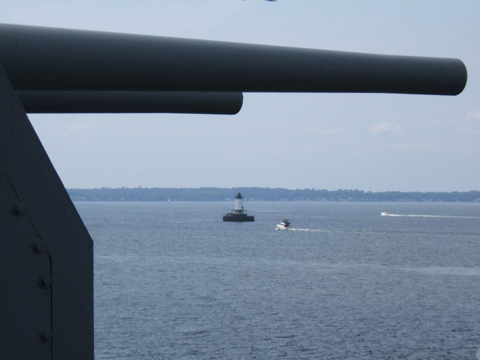Large gun barrel in foreground, lighthouse and boat on water in background