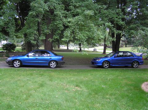 1996 Chevrolet Cavalier - Two blue sedans are parked on a driveway in a park-like setting with large trees.