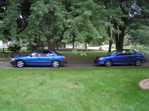 1996 Chevrolet Cavalier - Two blue sedans are parked side-by-side on a paved surface, with a grassy area and trees behind them.