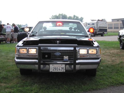 1991 Ford Crown Victoria - A police car with its hood open is parked on grass, with other vehicles and people in the background.