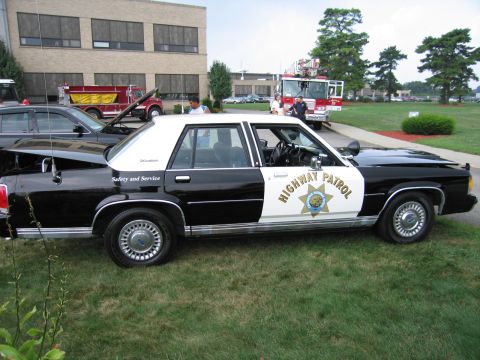 1991 Ford LTD Crown Victoria - black-and-white Highway Patrol cruiser with a gold star door shield sits on grass near a fire engine and building.