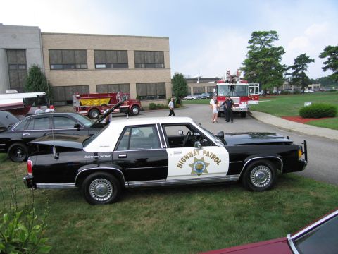 1991 Ford LTD Crown Victoria - black-and-white Highway Patrol sedan with star shield door markings parked on a lawn beside fire engines.