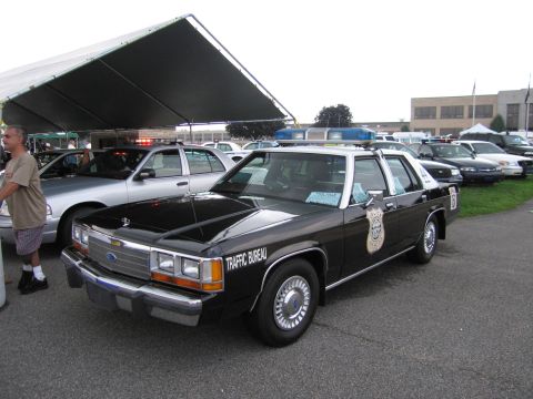 1991 Ford LTD Crown Victoria - black Ford LTD Crown Victoria marked Traffic Bureau with police shield and roof light bar parked under a tent.