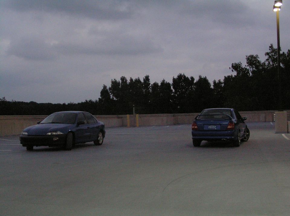 Two blue sedans parked on a rooftop parking lot under a cloudy sky