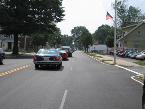 1991 Ford LTD Crown Victoria - dark sedan with headlights on drives up a sunny residential street lined with trees and a US flag.