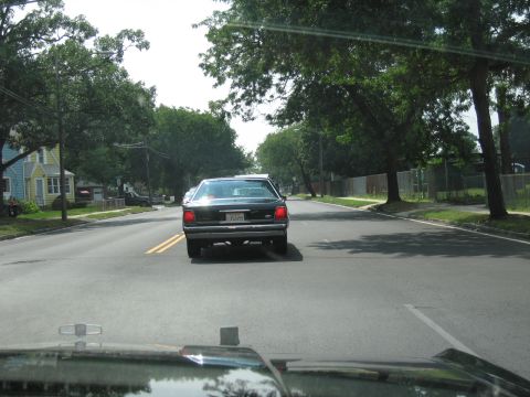 1991 Ford LTD Crown Victoria - Rear view through a windshield of a dark sedan driving down a green, tree-shaded residential street.