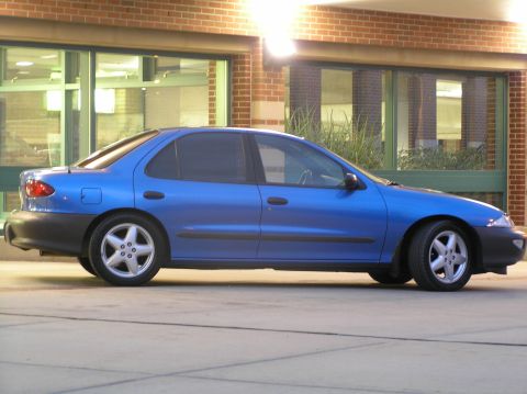 1996 Chevrolet Cavalier - A bright blue Chevrolet Cavalier four-door sedan shot in side profile at dusk on a concrete pad in front of a brick-and-glass building. It…