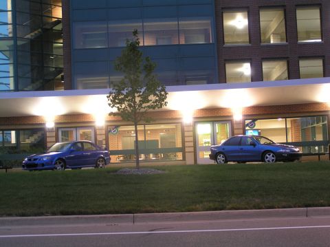 1996 Chevrolet Cavalier - Two blue sedans parked nose-to-nose at dusk along a lit brick and glass building, the left one a blue Mazda compact and the right a blue Ch…