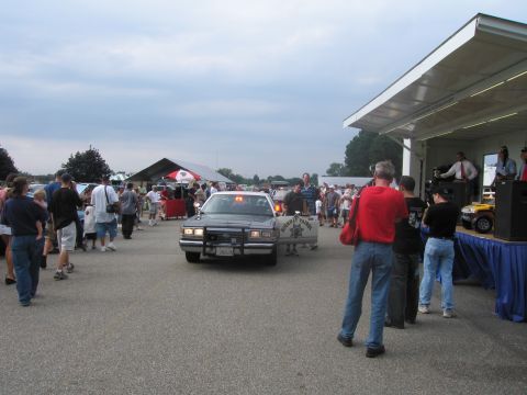 1991 Ford LTD Crown Victoria - black police sedan with amber lights on rolls through a crowd of spectators beside a covered event stage.