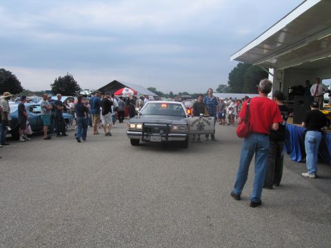 1991 Ford LTD Crown Victoria - black-and-white highway patrol sedan rolls through a busy outdoor car show as spectators gather under a tent and pavilion.
