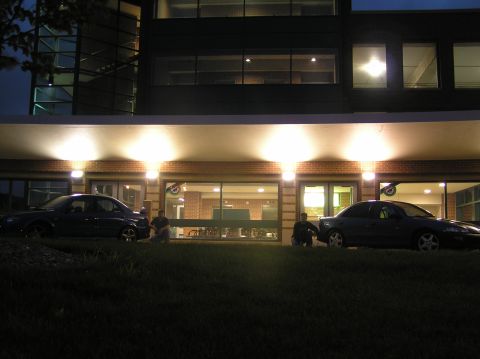 1996 Chevrolet Cavalier - Two sedans are parked in front of a lit building at dusk, with two individuals seated on the lawn.