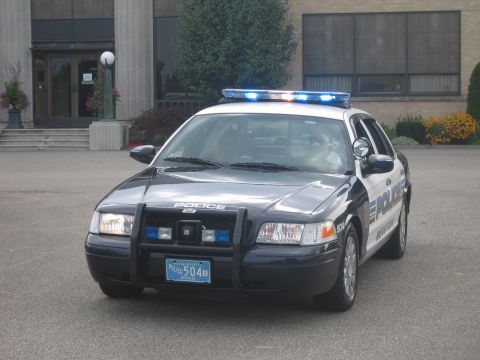 1991 Ford LTD Crown Victoria - Front three-quarter view of a dark Ford police cruiser marked POLICE with push bar and blue light bar on pavement.