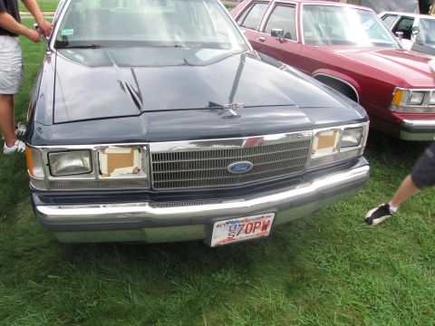1991 Ford LTD Crown Victoria - Front of a dark Ford full-size sedan with masked headlights and a Massachusetts US 70PW plate, people standing alongside.