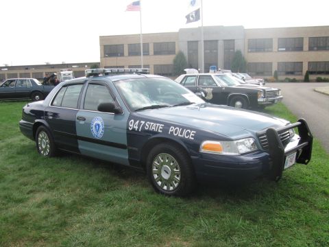 1991 Ford LTD Crown Victoria - blue-and-gray Ford cruiser marked 947 STATE POLICE with push bar parked on grass before a building flying flags.