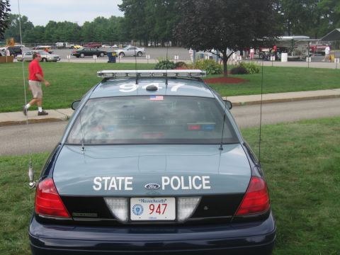 1991 Ford LTD Crown Victoria - Rear view of a blue-gray Ford State Police cruiser numbered 947 with roof light bar, parking lot and tent behind.