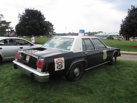 1991 Ford LTD Crown Victoria - Rear three-quarter view of a black-and-white Ford full-size police sedan with an Emergency 911 door shield on grass.