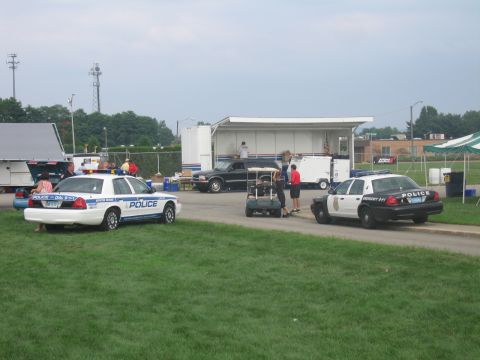 1991 Ford LTD Crown Victoria - Two white-and-blue marked police cruisers sit on grass at an outdoor show with vendor canopies and a trailer stage in the background.