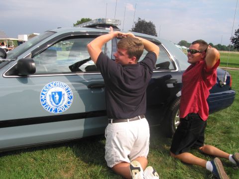 1991 Ford LTD Crown Victoria - Two young men kneel and strike poses beside a silver-blue Massachusetts State Police cruiser on a grassy field.