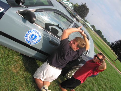 1991 Ford LTD Crown Victoria - young man strikes a flexing pose beside a Massachusetts State Police cruiser as another man laughs in the background on grass.