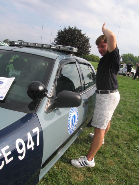 1991 Ford LTD Crown Victoria - young man in shorts raises an arm beside a silver-blue State Police cruiser marked 947, showing its light bar and door seal.