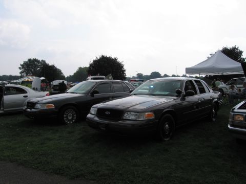 1991 Ford LTD Crown Victoria - Two dark gray Ford Crown Victoria-style sedans sit side by side on grass at an outdoor car show with tents behind them.