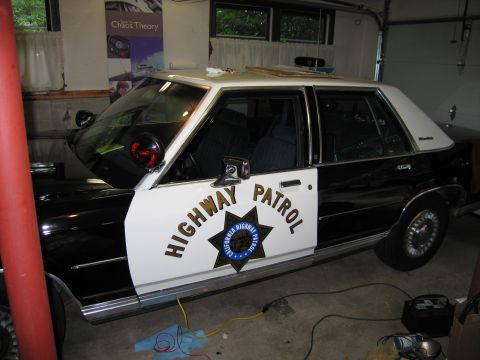 1991 Ford LTD Crown Victoria - Side profile of a black-and-white Highway Patrol sedan with a CHP door star and spotlight, parked in a cluttered garage.
