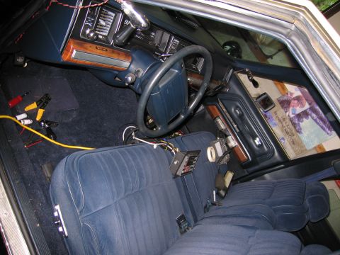 1991 Ford LTD Crown Victoria - Front interior of a sedan with blue bench seats and woodgrain dash, partly disassembled with wiring and yellow jumper cables visible.