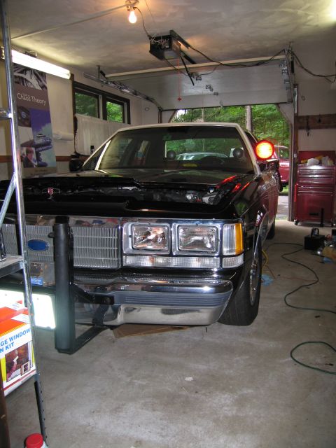 1991 Ford LTD Crown Victoria - Front of a dark Ford sedan with its hood propped open under garage lighting, dual rectangular headlights and chrome bumper visible.