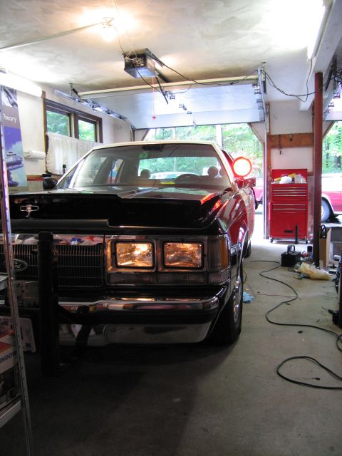 1991 Ford LTD Crown Victoria - Front of a dark Ford sedan with its headlights illuminated inside a garage, Ford grille emblem and chrome bumper visible.