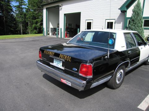 1991 Ford LTD Crown Victoria - Rear-quarter view of a black-and-white Ford Highway Patrol sedan parked on pavement in front of a building with open garage bays.
