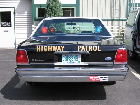 1991 Ford LTD Crown Victoria - Rear of a black Ford LTD Crown Victoria lettered Highway Patrol with NH plate CHP8849, parked outside a building beside a van.