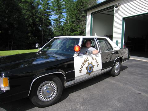 1991 Ford LTD Crown Victoria - smiling man sits in a black-and-white police sedan marked HIGHWAY PATROL with a star emblem, door open beside an open garage bay.