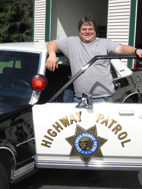 1991 Ford LTD Crown Victoria - man in a gray shirt leans on the front of a black-and-white police car displaying the California Highway Patrol star and red spotlight.