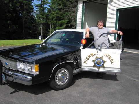 1991 Ford LTD Crown Victoria - man stands at the open driver door of a black-and-white Ford police sedan marked HIGHWAY PATROL, parked before a garage.