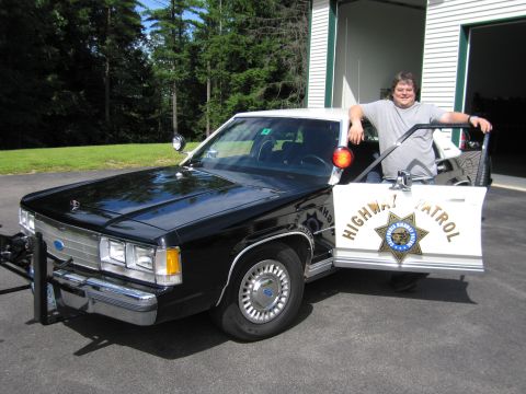 1991 Ford LTD Crown Victoria - Front three-quarter view of a black-and-white Ford police sedan with CHP star emblem and red spotlight, a man standing at the open door.